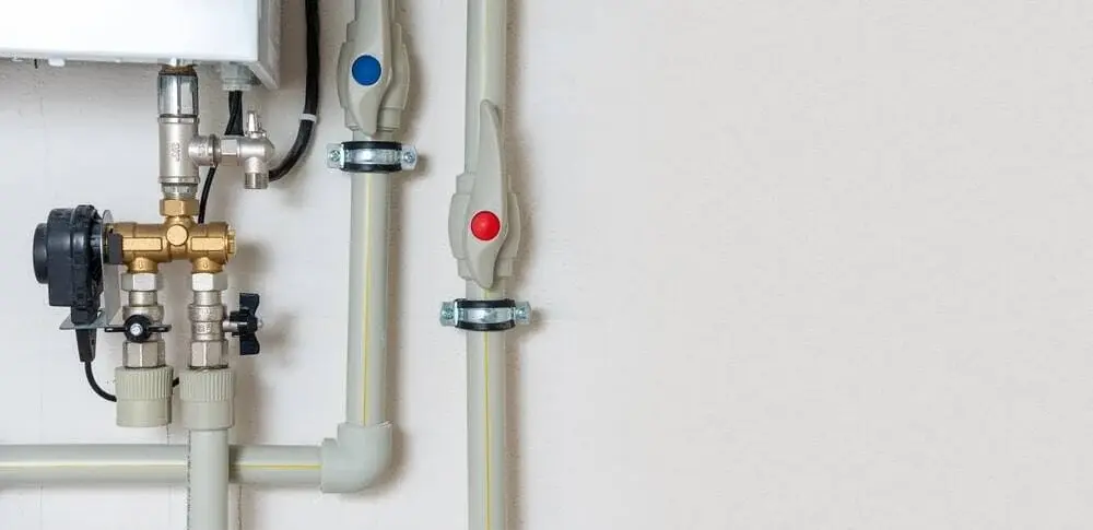 A detailed close-up photograph of newly installed white PEX boiler pipework on a clean wall. The left side shows complex brass fittings and a dark grey motorized zone valve connecting to a boiler. To the right, two main flow-and-return pipes are visible. One features a grey ball-valve handle with a blue indicator, and the other is a gate-valve with a red indicator. The pipes are secured by metal clamps with rubber liners. There is an open space on the right, perfect for text overlays or to show the contrast with previous, perhaps corroded, pipes. The work looks professional and clean, indicating the high standard of a quality plumber in Luton.