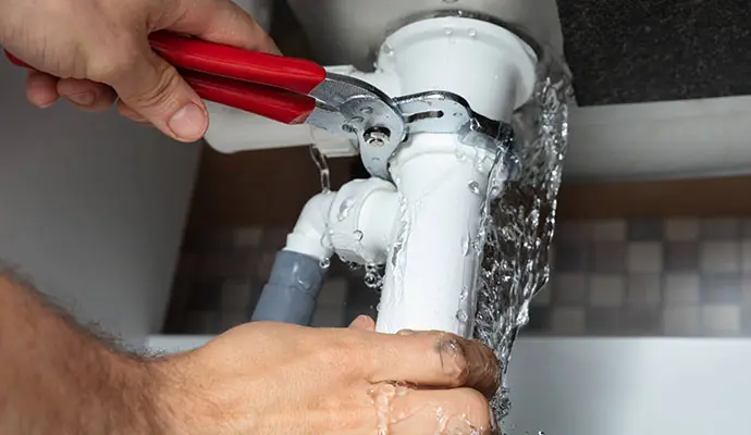 An emergency plumber in Luton using a pipe wrench to fix a spraying water leak on a white plastic sink P-trap trap under a cabinet.